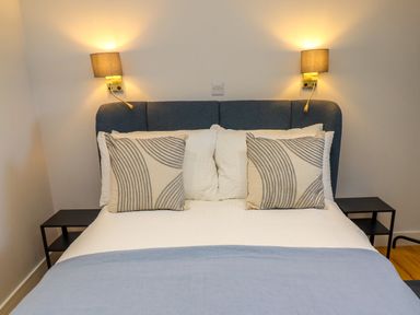 A bed with blue headboard white sheets and patterned pillows with bedside tables and wall lamps at Ocean View in Alnmouth