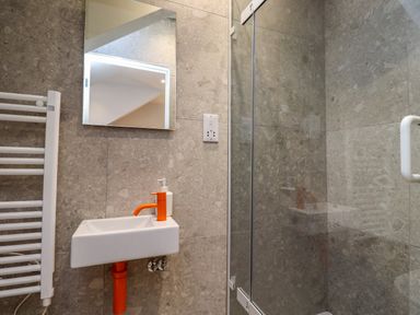 A bathroom with a small sink with orange faucet and pipe a wall mirror a white towel radiator and a glass shower door at Ocean View in Alnmouth