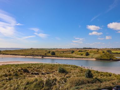 A river bordered by sandy banks and grassy hills with people near the water under a blue sky at Ocean View in Alnmouth