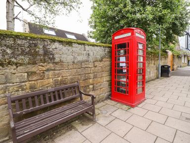 A brown bench next to a red telephone booth on a paved sidewalk by a stone wall with a tree behind at Ocean View in Alnmouth