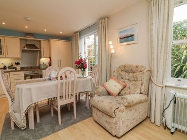 A dining area with a table set with a tablecloth and flowers next to a kitchen and a floral armchair at Archie's in Minehead