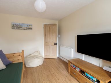 A room with a wooden door, bean bag, wood floor, sofa with cushions, wall-mounted TV, and wooden TV stand with board games at Beach House in Winterton-On-Sea