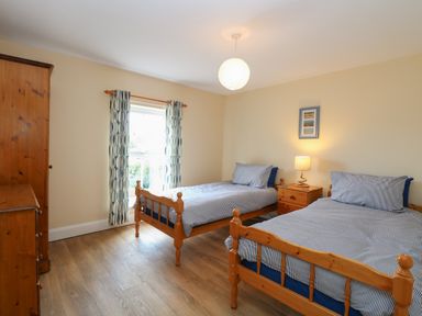 A bedroom with two single wooden beds a wooden side table with lamp a wooden wardrobe a window with patterned curtains and wooden floor at Beach House in Winterton-On-Sea