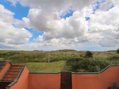 A garden with a lawn and greenery beyond a red wall and wooden gate under a cloudy sky at Beach House in Winterton-On-Sea