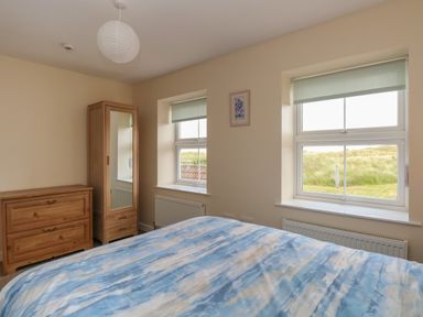 A bedroom with a bed with blue and white bedding a wooden dresser a wooden wardrobe with mirror two windows with roller blinds at Beach House in Winterton-On-Sea