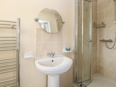 A bathroom with a pedestal sink a round mirror a glass shower enclosure and a chrome towel radiator at Beach House in Winterton-On-Sea