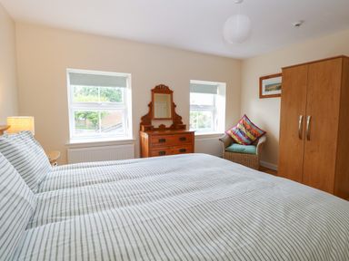 A bedroom with a bed striped bedding a wooden dresser a wicker chair with colorful cushions and a wooden wardrobe at Beach House in Winterton-On-Sea
