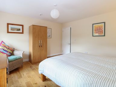 A bedroom with a striped bed, wooden wardrobe, wicker chair with colorful cushions, wooden floor and framed pictures on walls at Beach House in Winterton-On-Sea