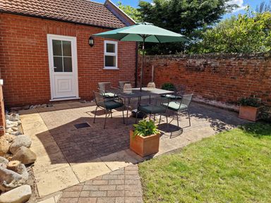 A patio with a table and six chairs under an umbrella next to a brick house and a brick wall with plants in pots at Beach House in Winterton-On-Sea