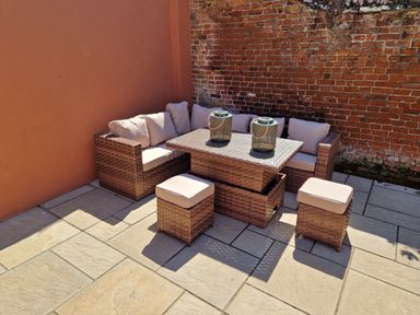 An outdoor seating area with a wicker corner sofa a table and two stools on a tiled patio between a brick wall and a stucco wall at Beach House in Winterton-On-Sea