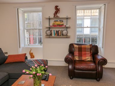 A living room with a brown leather armchair, a grey sofa, a wooden table with pink flowers, and shelves with books and games at The Old Factory House in Berwick-Upon-Tweed