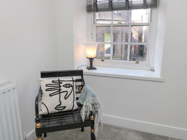 A corner with a black wooden chair holding a cushion and blanket next to a window with a lamp on the windowsill at The Old Factory House in Berwick-Upon-Tweed