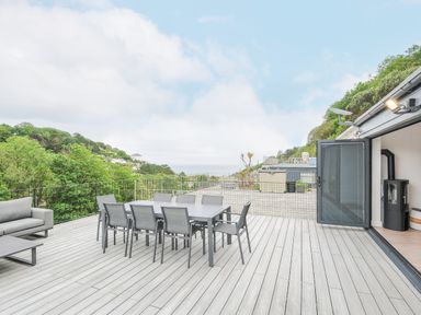 An outdoor terrace with a dining table and chairs a sofa and a coffee table overlooking trees and hills at 100 Hillside Villas in Looe