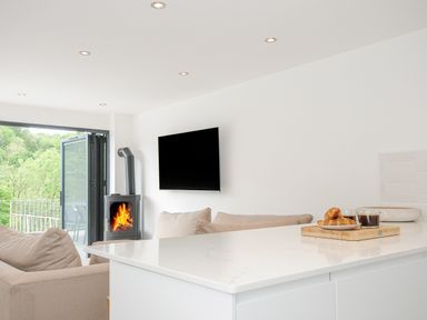 A living room with sofas a wood-burning stove a wall-mounted TV and a kitchen island with coffee and pastries on a tray at 100 Hillside Villas in Looe