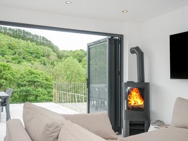 A living room with a beige sofa a black wood stove with fire a wall-mounted TV and an open glass door leading to a balcony with outdoor furniture and a view of green trees at 100 Hillside Villas in Looe