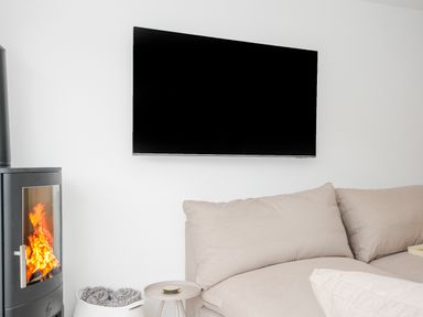 A living area with a wood burning stove a flat screen tv mounted on a white wall a beige sofa a small round side table and a basket with textiles at 100 Hillside Villas in Looe