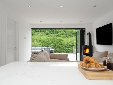 A modern living room with beige sofas a wood burning stove a wall mounted TV and an open patio door showing outdoor furniture and green trees at 100 Hillside Villas in Looe