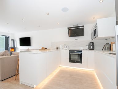 A kitchen with white cabinets a stove oven microwave and a toaster next to a living area with sofas and a wall-mounted TV at 100 Hillside Villas in Looe