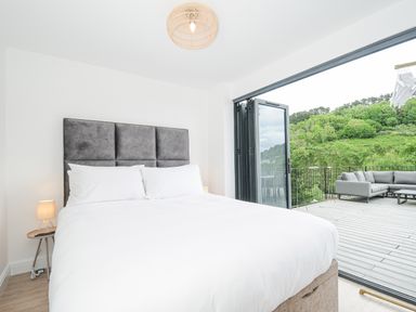 A bedroom with a large bed next to an open balcony door showing outdoor seating and greenery at 100 Hillside Villas in Looe