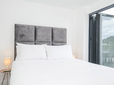 A bedroom with a bed with white bedding and pillows a grey padded headboard a bedside table with a lamp and a folding glass door leading outside at 100 Hillside Villas in Looe