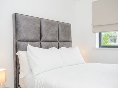 A bed with a gray padded headboard white pillows and white bedding near a window with a beige blind at 100 Hillside Villas in Looe