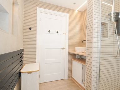 A bathroom with a white door black hooks a white laundry basket a black towel radiator a sink on a wooden countertop and a glass shower enclosure at 100 Hillside Villas in Looe