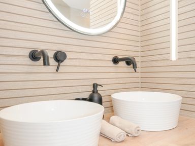 A bathroom sink area with two white vessel sinks black faucets a round mirror and beige textured wall panels at 100 Hillside Villas in Looe