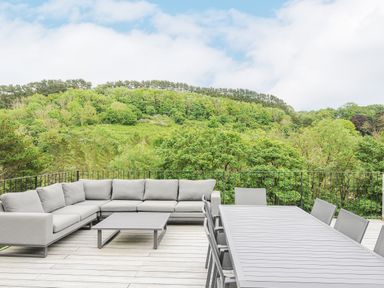 An outdoor patio with grey sofa and chairs around tables overlooking a green forest at 100 Hillside Villas in Looe