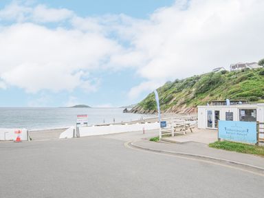 A beach with a resort building and picnic tables near a hillside with houses at Millendreath Beach Resort in Looe