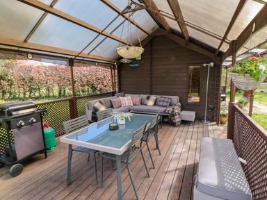 A covered outdoor patio with a dining table and chairs a sofa with cushions a barbecue and hanging plants at Snape Castle Garden Lodge Snape near Bedale