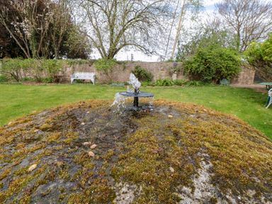A garden with a moss-covered stone water fountain and greenery by a stone wall at Snape Castle Garden Lodge Snape near Bedale