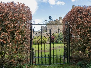 An iron wrought gate between trimmed bushes with a castle and garden in the background at Snape Castle Garden Lodge in Snape near Bedale