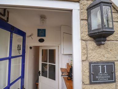 An open blue door leading to a narrow hallway with a white door, a sign on the wall, and a stone exterior wall with a lantern and a plaque reading The Old Ropemakers Cottage at Bridport
