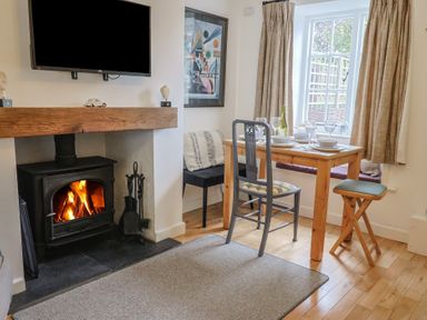 A dining area with a wooden table set for four near a window and a fireplace with a fire burning at Old Ropemakers Cottage in Bridport