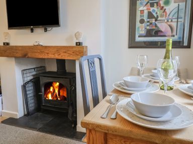 A dining table set with plates bowls wine glasses and a bottle near a fireplace with a fire burning in Old Ropemakers Cottage in Bridport