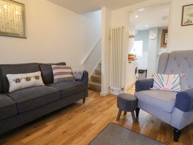 A living room with a dark gray sofa with cushions, a light blue armchair with a cushion, a small stool, wooden floor, stairs, and a view into a kitchen at Old Ropemakers Cottage in Bridport