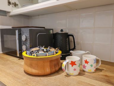 A kitchen countertop with a microwave kettle wooden bowl with snacks and two star patterned mugs at Old Ropemakers Cottage in Bridport