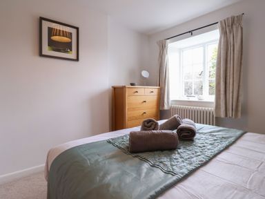 A bedroom with a bed covered in white and teal linens with rolled towels on it and a wooden dresser near a window with curtains at Old Ropemakers Cottage in Bridport