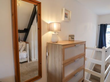 A wooden chest of drawers with a lamp and container on top next to a full-length mirror showing a bed and wooden beams at Old Ropemakers Cottage in Bridport