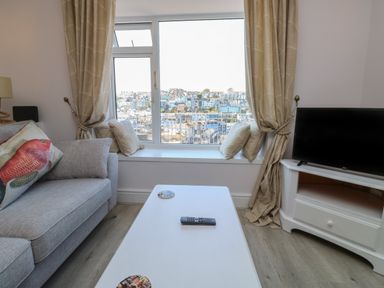A living room with a grey sofa with fish pillow a white coffee table with remote and coasters TV on white cabinet and large window with cushions and beige curtains at Fishnets in Brixham