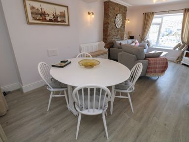 A dining area with a round white table and four white chairs next to a living room with grey sofas and a large window at Fishnets in Brixham