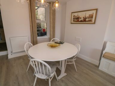 A dining area with a round white table and four chairs a window with beige curtains and a painting on the wall at Fishnets in Brixham