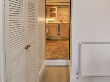 A hallway with white louvered doors and a radiator leading to a kitchen with wooden cabinets at Fishnets in Brixham