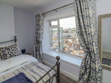 A bedroom with a metal bedframe next to a window with patterned curtains and a view of a town at Fishnets in Brixham