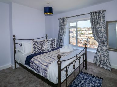 A bedroom with a metal bed, patterned cushions, curtains, and a window overlooking a town at Fishnets in Brixham