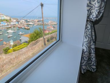 A window with patterned curtains showing a view of a harbor with boats and buildings at Fishnets in Brixham