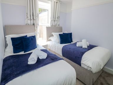 A bedroom with two single beds with blue cushions and throws a window with curtains between them at Fishnets in Brixham