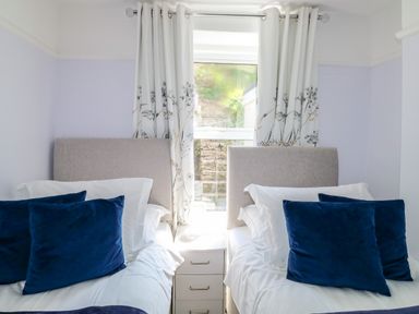 A bedroom with two single beds blue pillows and a bedside table under a window with floral curtains at Fishnets in Brixham