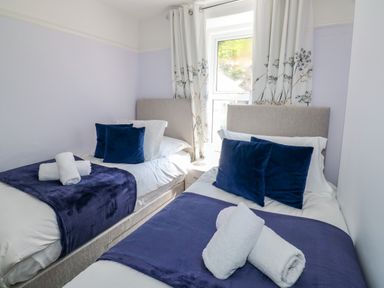 A bedroom with two single beds with blue throws and pillows and white rolled towels in front of a window with floral curtains at Fishnets in Brixham