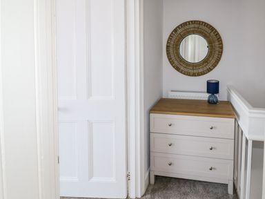 A white chest of drawers with a wooden top a blue lamp and a round decorative wall mirror above at Fishnets in Brixham